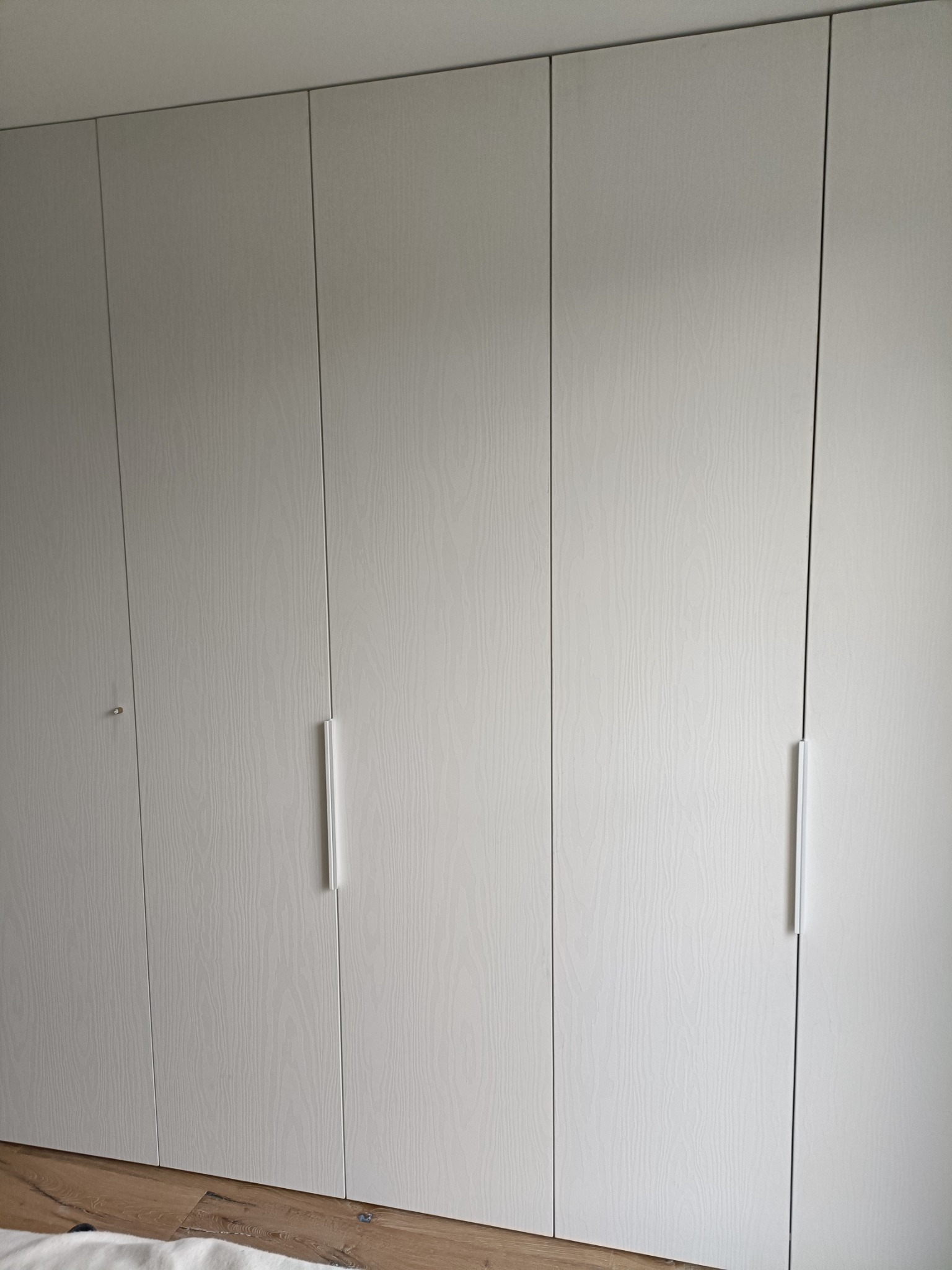 Biała szafa wnękowa z minimalistycznymi, długimi uchwytami i widoczną strukturą drewna na frontach, sięgająca od podłogi do sufitu.