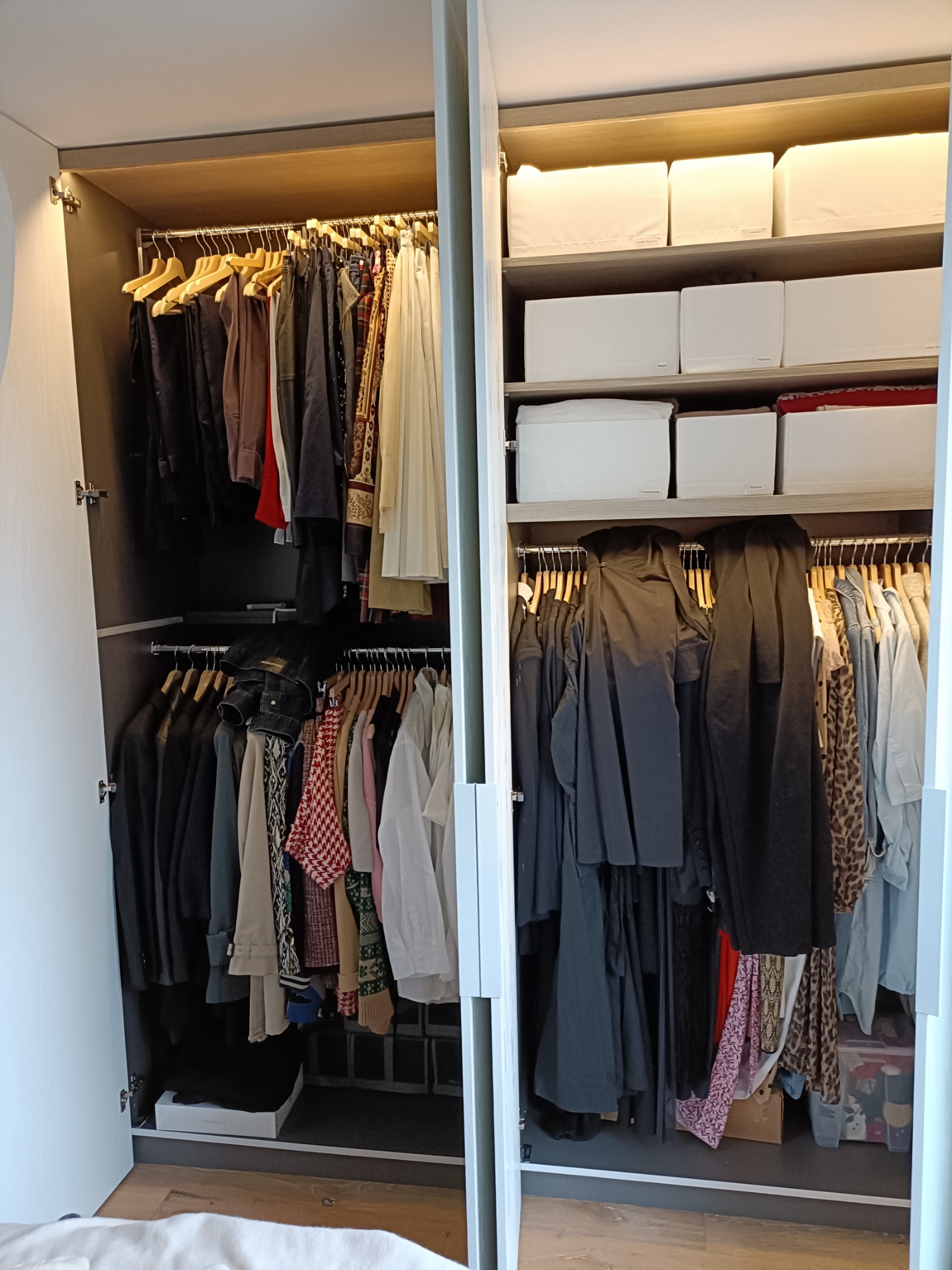 Wnętrze szafy wnękowej z otwartymi drzwiami, widoczne ubrania wiszące na wieszakach i białe pudełka do przechowywania na półkach.