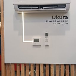 Hydrovolt Serwis - Biała klimatyzacja Ukura z panelem sterowania i pilotem na jasnym tle, otoczona drewnianą konstrukcją. Widoczne parametry techniczne urządzenia.