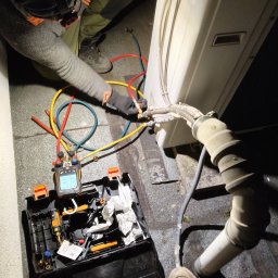 Hydrovolt Serwis - Serwisant w trakcie podłączania przewodów do klimatyzatora zewnętrznego. Widoczny manometr cyfrowy i narzędzia w otwartej skrzynce. Praca w słabym oświetleniu.