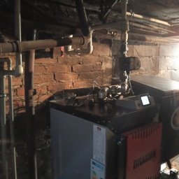 Hydrovolt Serwis - Kocioł na paliwo stałe w piwnicy z instalacją hydrauliczną. Widoczne rury, ceglana ściana i etykieta energetyczna na urządzeniu. Oświetlenie punktowe.