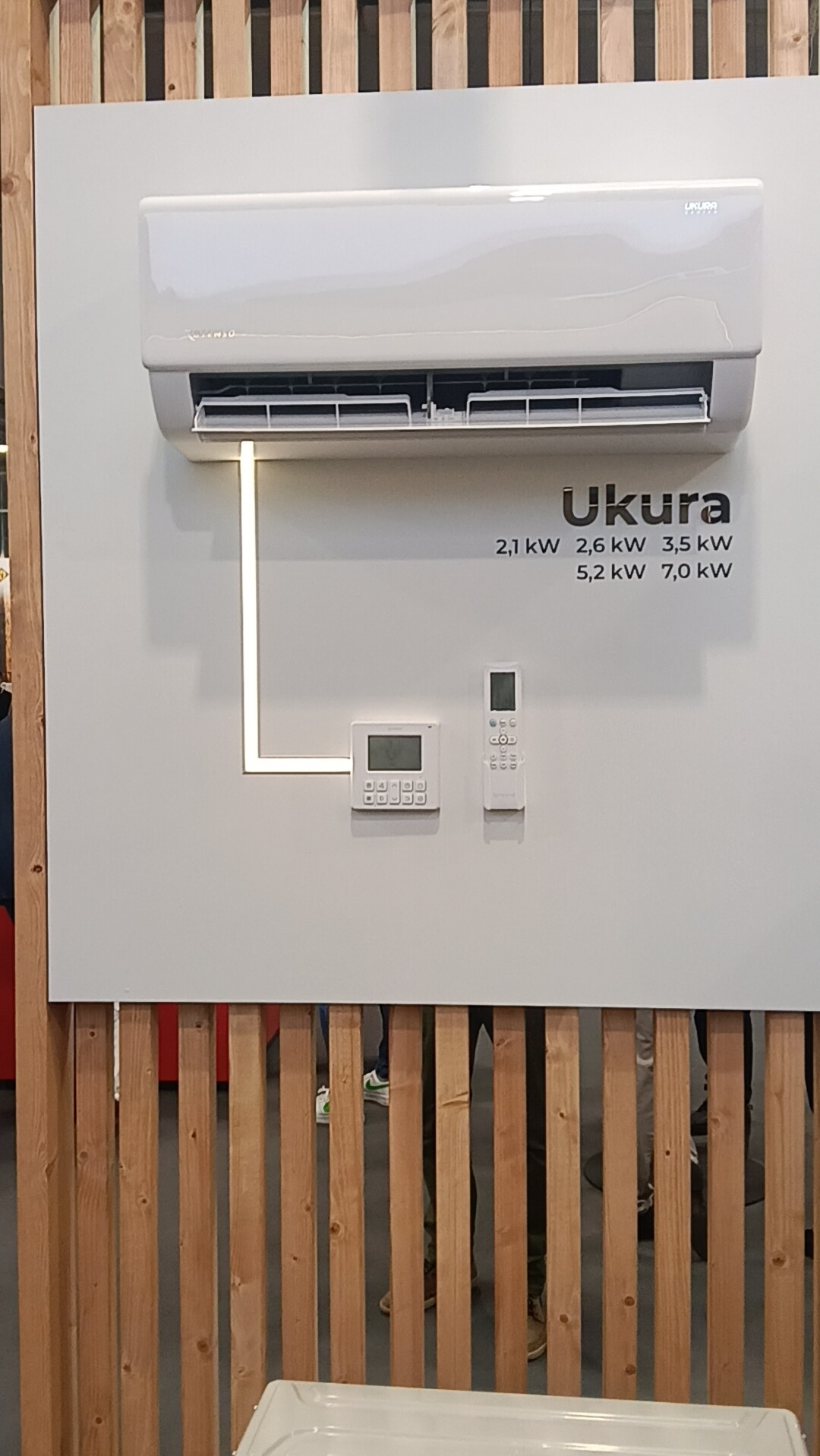 Biała klimatyzacja Ukura z panelem sterowania i pilotem na jasnym tle, otoczona drewnianą konstrukcją. Widoczne parametry techniczne urządzenia.