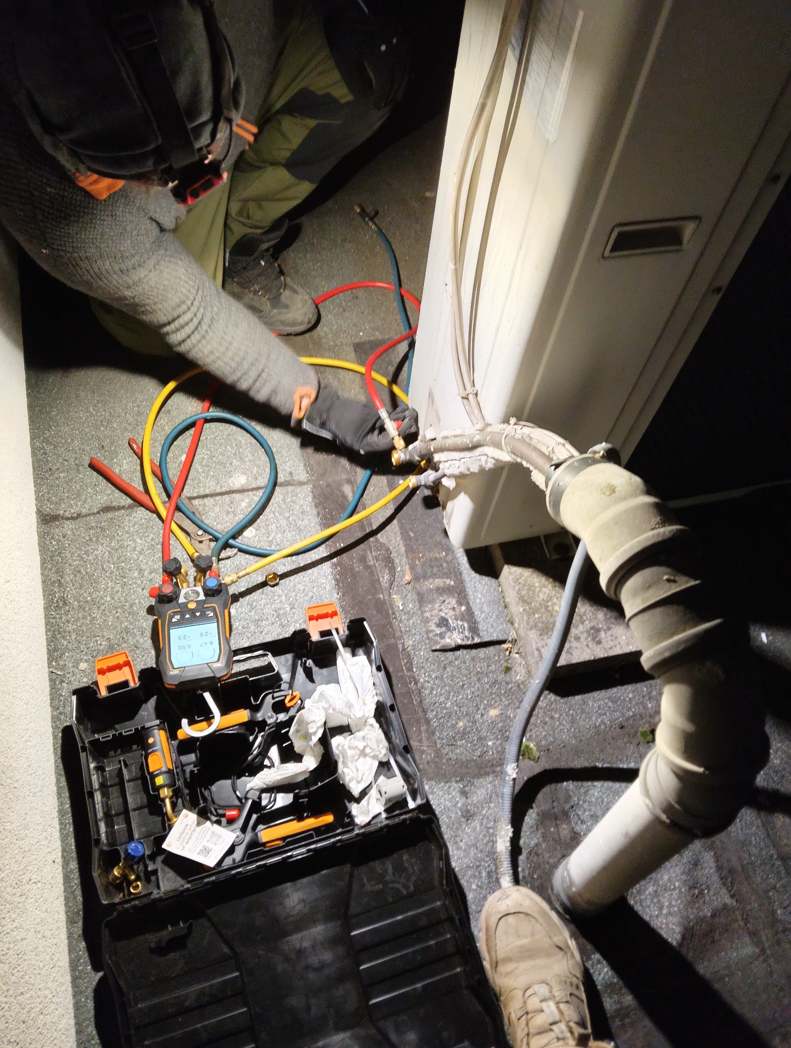 Serwisant w trakcie podłączania przewodów do klimatyzatora zewnętrznego. Widoczny manometr cyfrowy i narzędzia w otwartej skrzynce. Praca w słabym oświetleniu.