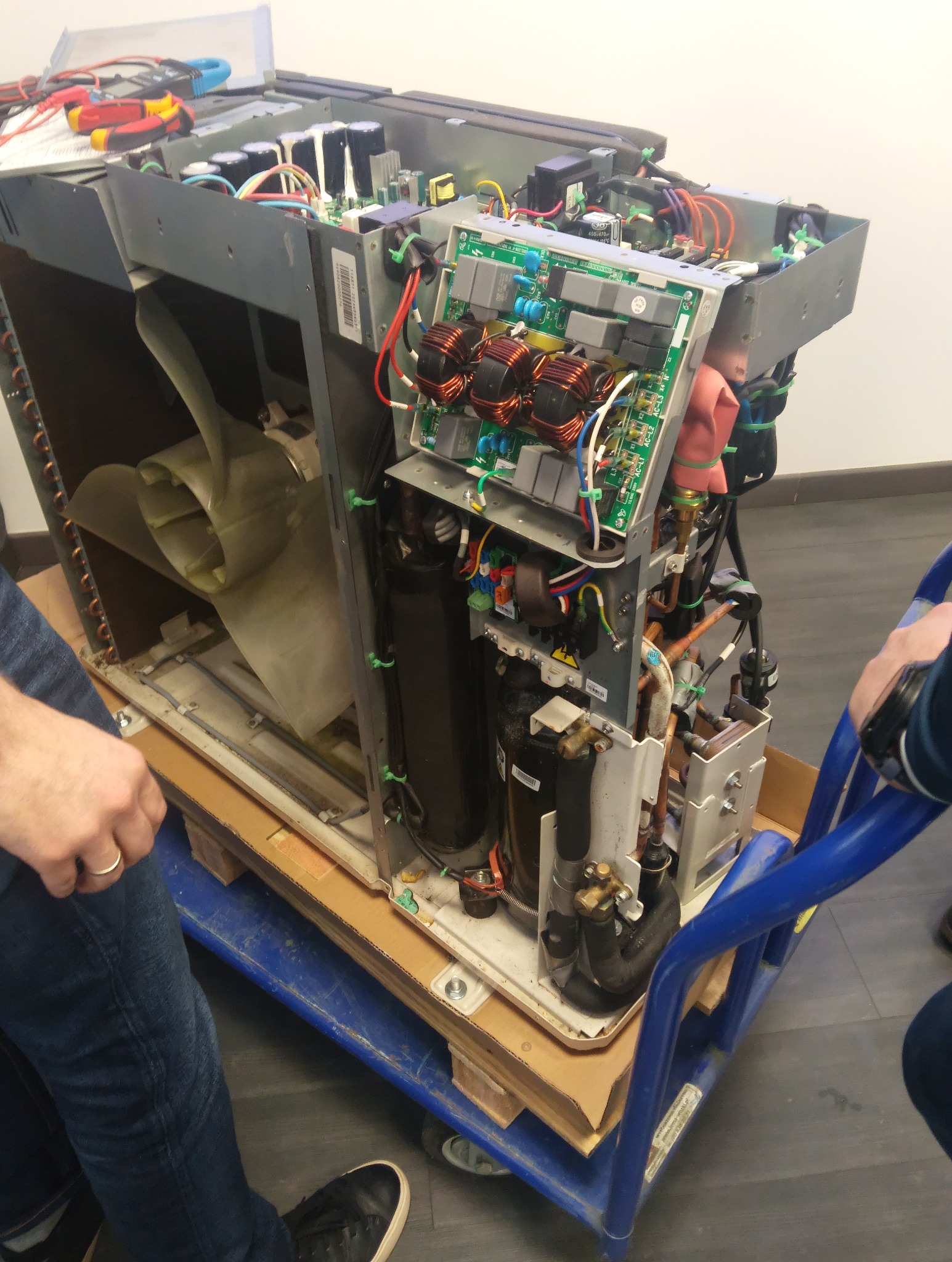 Otwarta obudowa klimatyzatora na wózku transportowym, widoczne elementy wewnętrzne: wentylator, sprężarka, elektronika. Serwis klimatyzacji w trakcie naprawy.