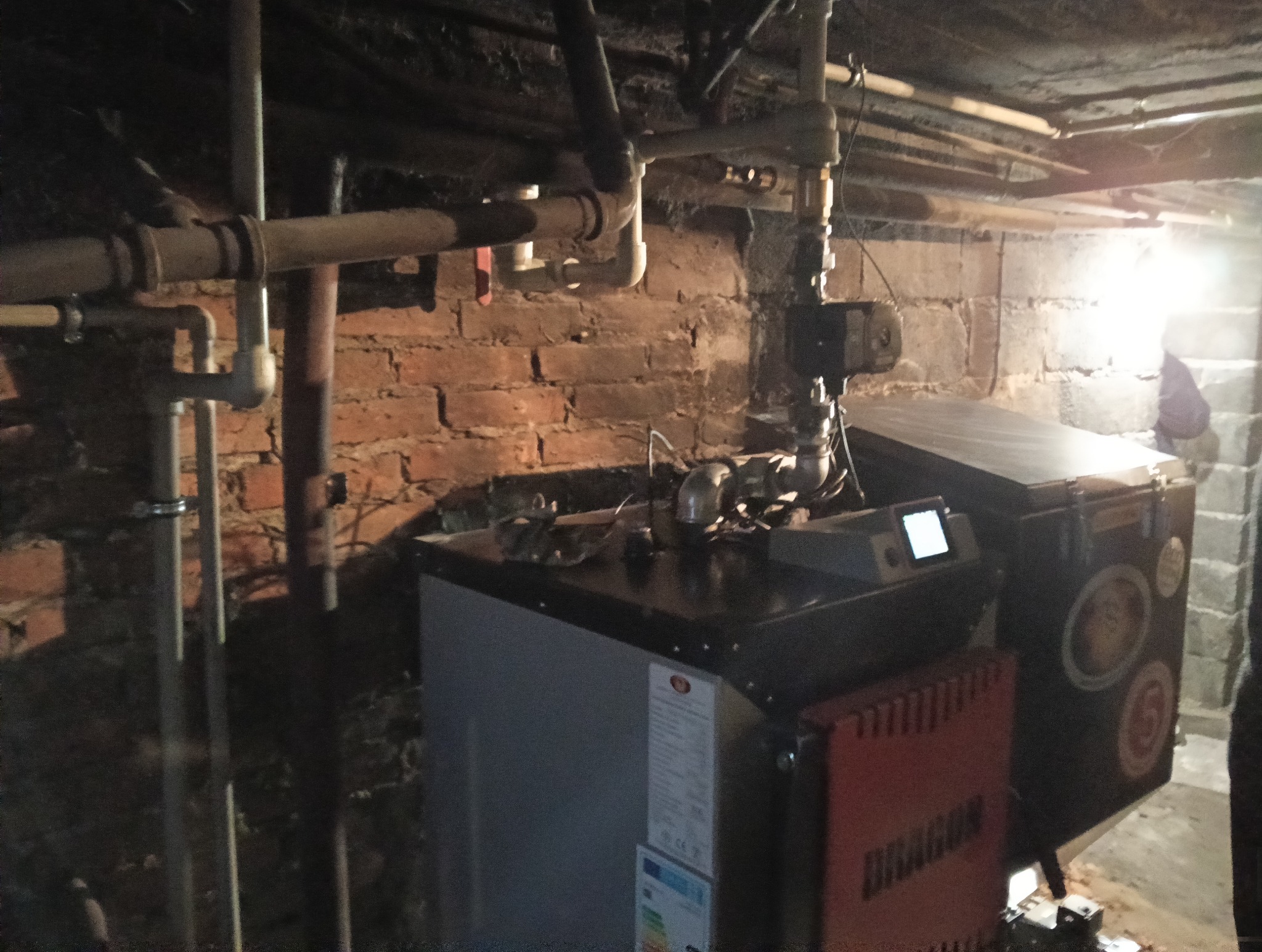 Kocioł na paliwo stałe w piwnicy z instalacją hydrauliczną. Widoczne rury, ceglana ściana i etykieta energetyczna na urządzeniu. Oświetlenie punktowe.