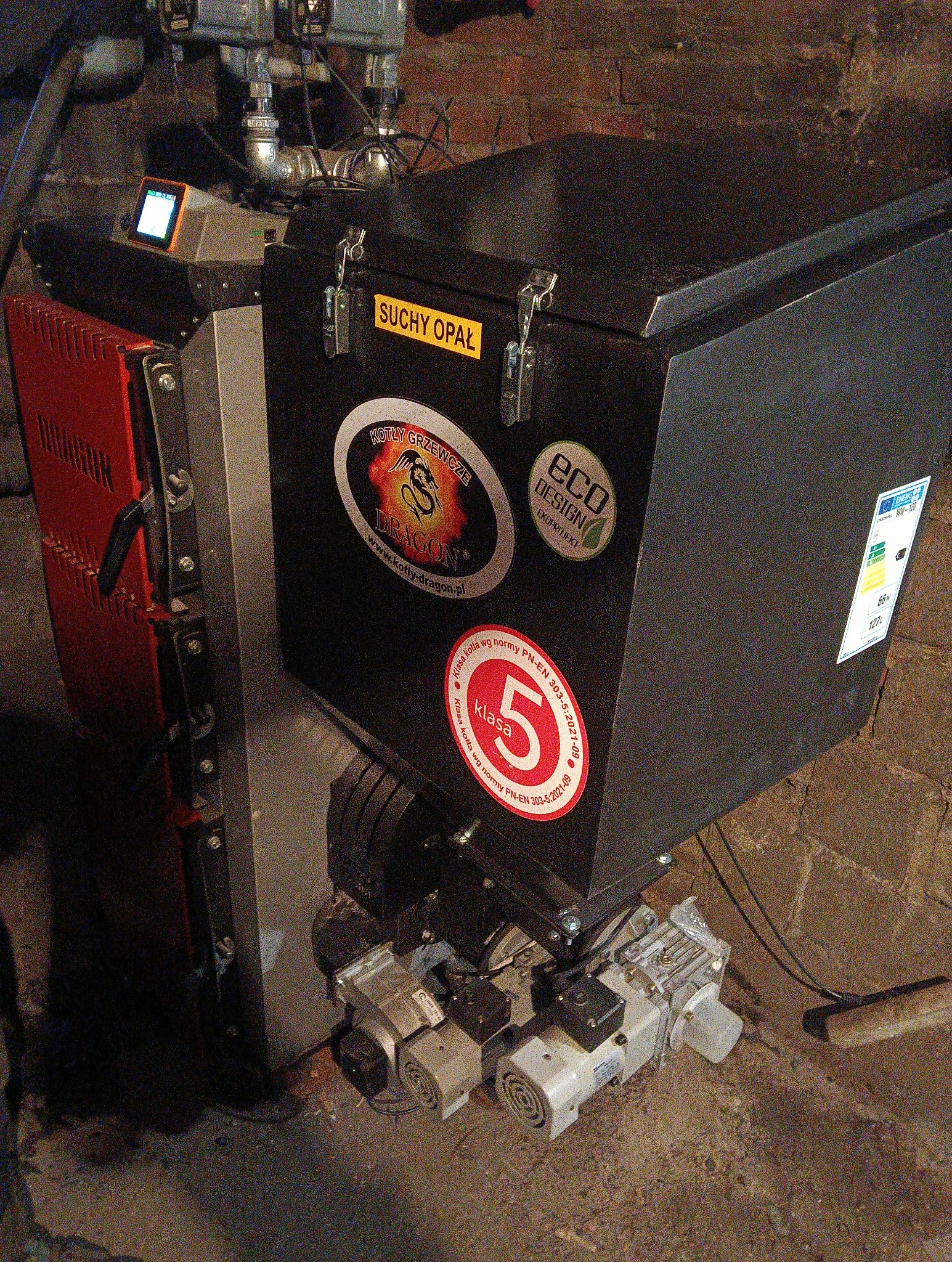 Kocioł grzewczy na paliwo stałe z oznaczeniami klasy 5 i EcoDesign, widoczny w kotłowni z ceglaną ścianą. Na kotle naklejki informacyjne i logo producenta.