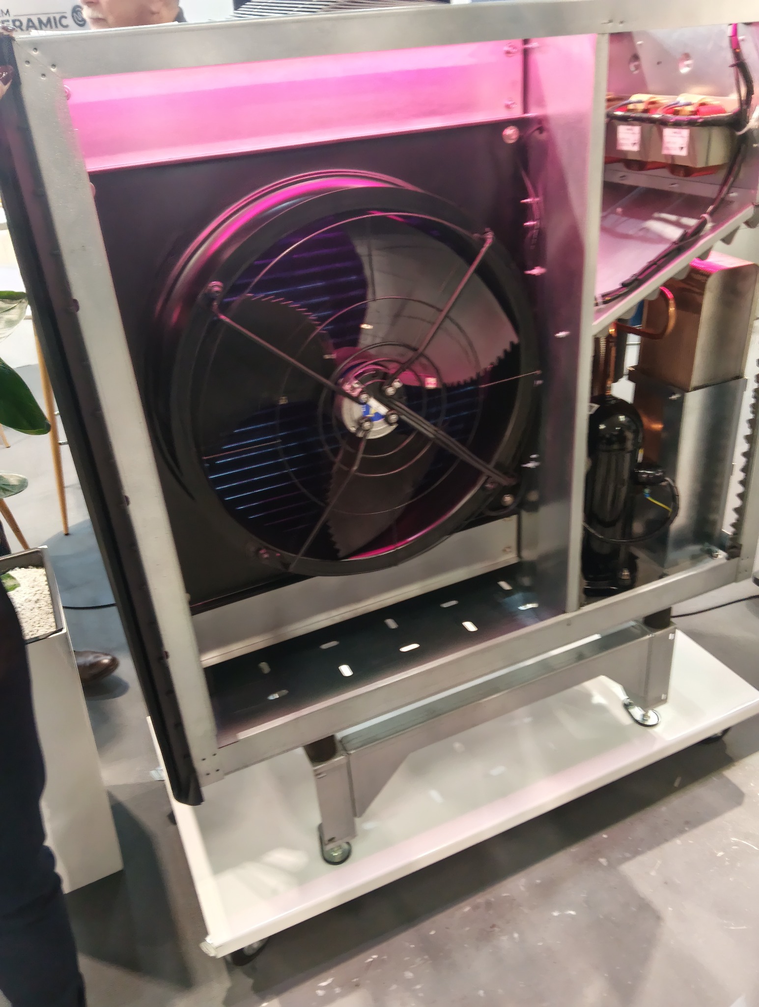 Wnętrze metalowej obudowy z wentylatorem i elementami instalacji, oświetlone różowym światłem. Widoczne przewody i podzespoły. Urządzenie na kółkach.