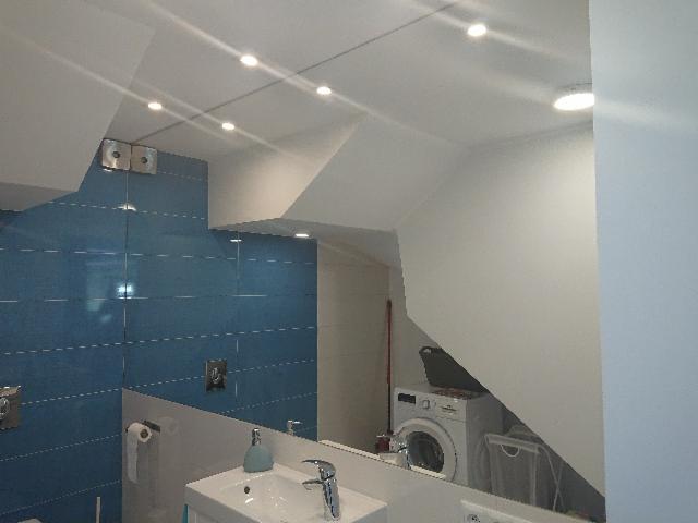 Nowoczesna łazienka z niebieskimi szklanymi panelami ściennymi, mocowanymi za pomocą metalowych uchwytów punktowych, odbijającymi w lustrze pralkę.