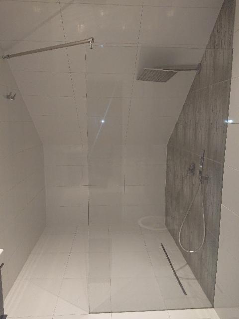 Nowoczesna kabina prysznicowa ze szklaną ścianką, deszczownicą i odpływem liniowym w minimalistycznej łazience z białymi płytkami i akcentem szarej ściany.