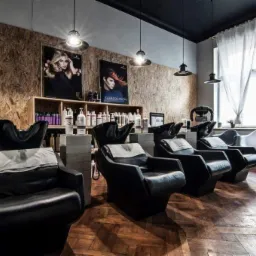 Wnętrze salonu fryzjerskiego z czterema czarnymi fotelami z umywalkami, drewniana ściana z plakatami i półkami z kosmetykami, wiszące lampy i podłoga z parkietu.