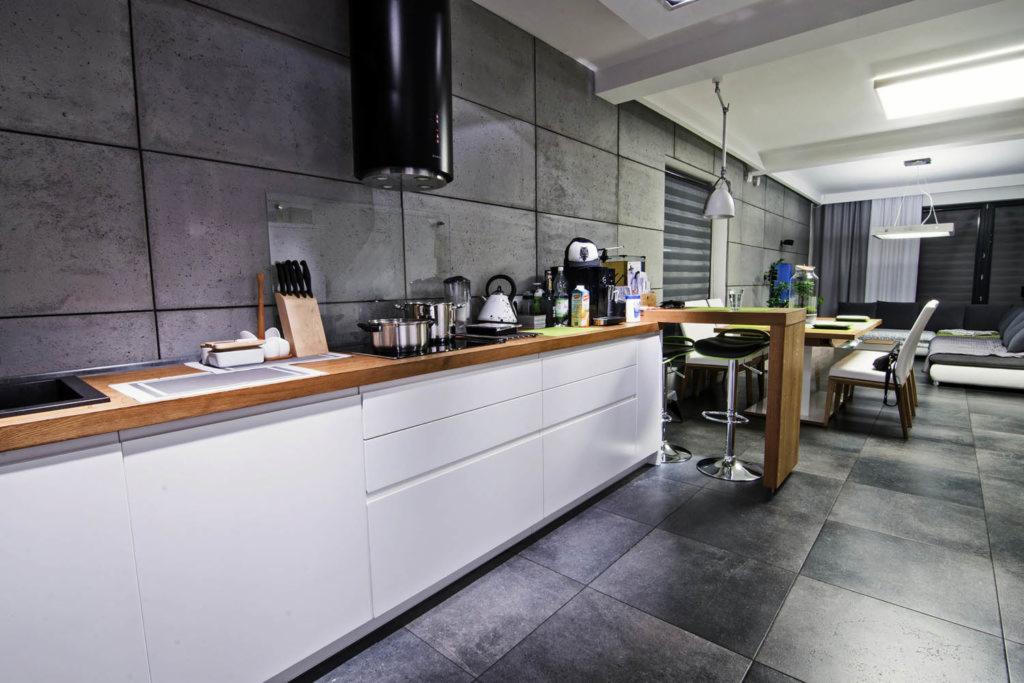 Nowoczesna kuchnia z szarymi płytami na ścianie, białymi szafkami i drewnianym blatem, przechodząca w jadalnię z wysokimi krzesłami barowymi i widokiem na salon z szarą sofą.