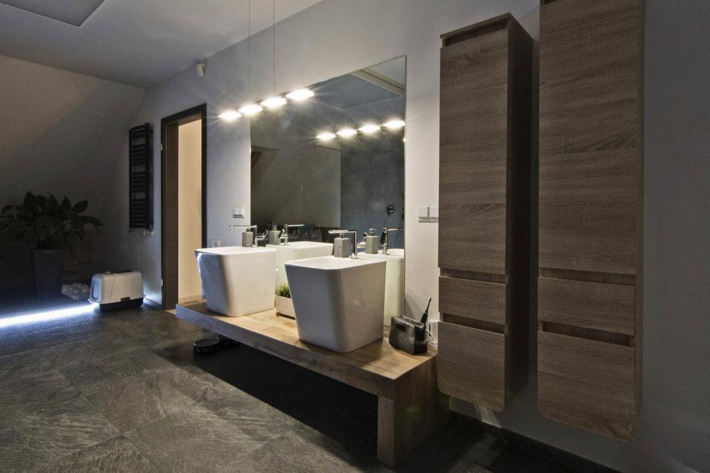 Nowoczesna łazienka w domu jednorodzinnym: dwie kwadratowe umywalki nablatowe z chromowanymi bateriami, duże lustro z oświetleniem LED, wysokie szafki w kolorze drewna, szara podłoga z płytek...