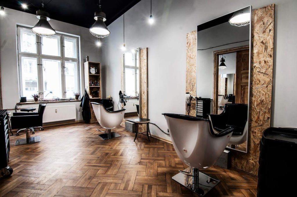 Wnętrze salonu fryzjerskiego w stylu loftowym z parkietem w jodełkę, białymi ścianami, dużymi lustrami obramowanymi płytami OSB i nowoczesnymi fotelami fryzjerskimi w kształcie muszli.