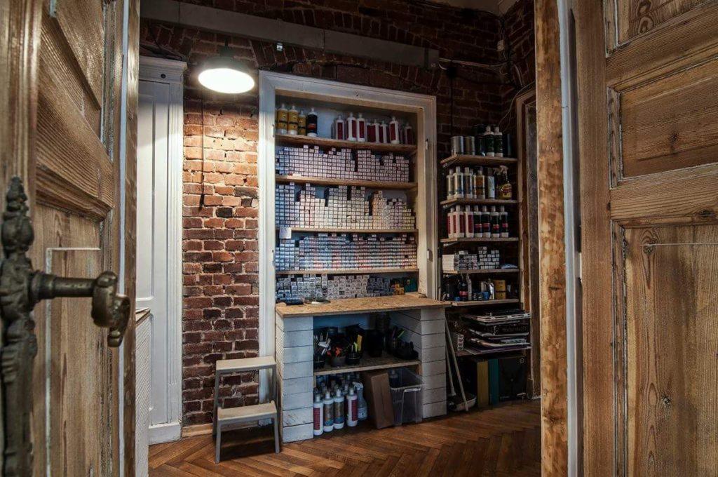 Wnętrze salonu fryzjerskiego w stylu loftowym: Półki z farbami do włosów i kosmetykami na tle ceglanej ściany, widoczne przez otwarte drewniane drzwi z ozdobnym uchwytem.