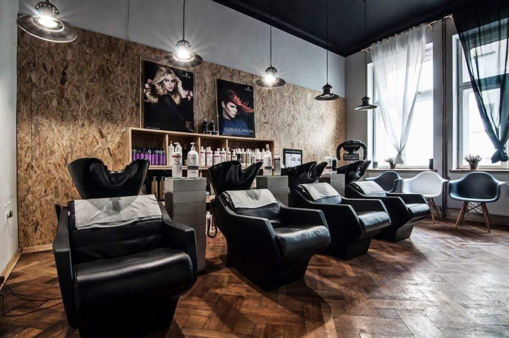Wnętrze salonu fryzjerskiego z czterema czarnymi fotelami z umywalkami, drewniana ściana z plakatami i półkami z kosmetykami, wiszące lampy i podłoga z parkietu.