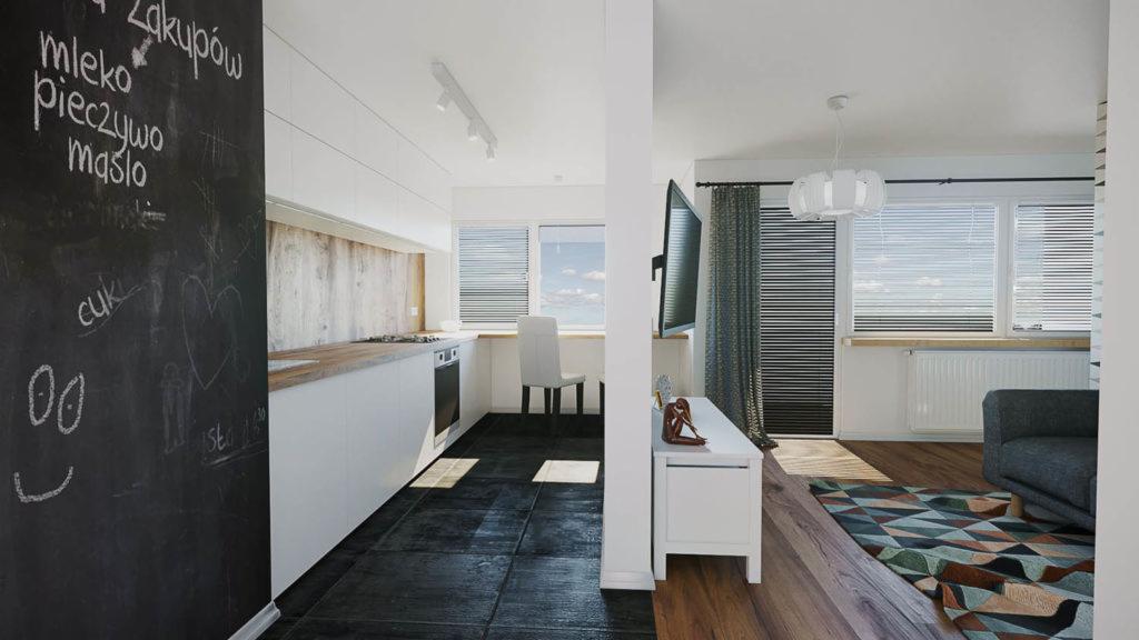 Wnętrze kawalerki z aneksem kuchennym, czarna ściana z rysunkami kredą, białe meble kuchenne, czarna podłoga z płytek, widok na salon z szarą sofą i kolorowym dywanem.