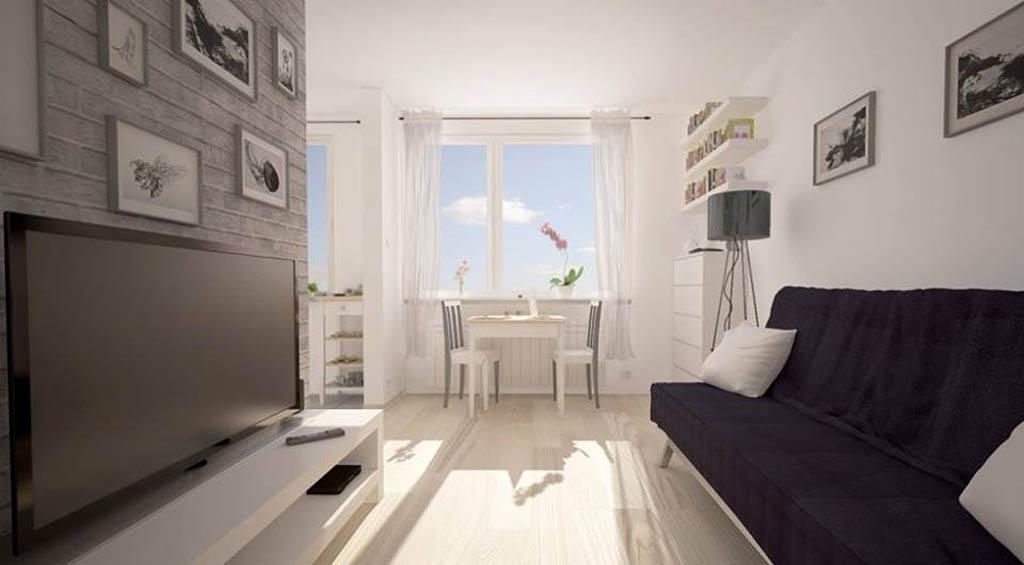 Wnętrze kawalerki z szarą ceglaną ścianą, telewizorem na białej szafce RTV, ciemną sofą, białym stolikiem z krzesłami przy oknie i drewnianą podłogą.