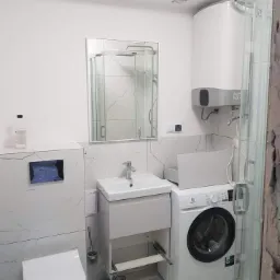 Biała łazienka z kabiną prysznicową, umywalką z szafką z wysuniętą szufladą, pralką Electrolux i bojlerem. Widoczne płytki imitujące marmur na ścianach i podłodze.