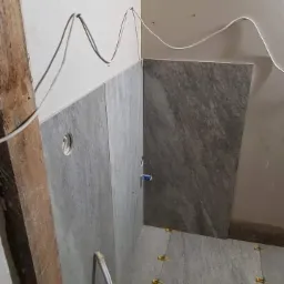 Szare płytki imitujące kamień na ścianie i podłodze w trakcie montażu, widoczne żółte krzyżyki dystansowe i wystające przewody elektryczne, drewniana listwa przypodłogowa w lewym rogu.