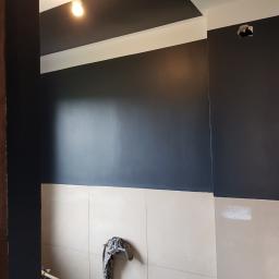 Krystiano - Świeżo pomalowana na ciemny kolor ściana w łazience, z widocznymi jasnymi płytkami ceramicznymi na dole, niedomalowanym fragmentem ściany i wiszącą szmatką, oraz widocznymi narzędziami malarskimi.