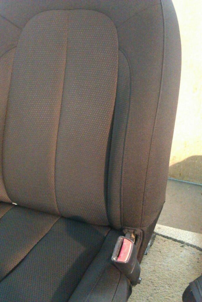 Fragment tapicerki samochodowej w kolorze ciemnoszarym z widocznym zapięciem pasa bezpieczeństwa i delikatną fakturą materiału.