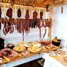 Stół cateringowy z wędlinami, serami i pieczywem pod drewnianą konstrukcją z wiszącymi kiełbasami i serami w stylu rustykalnym, dekoracja z dyni w tle.