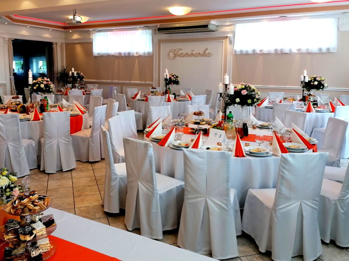 Uroczysta sala bankietowa z okrągłymi stołami nakrytymi białymi obrusami i pomarańczowymi akcentami, krzesła w białych pokrowcach, na stołach kompozycje kwiatowe i świeczniki, w tle dekoracyjny...