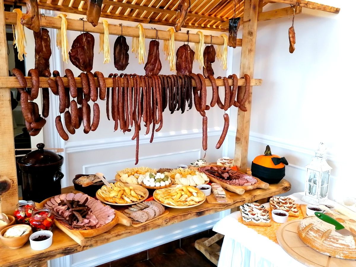Stół cateringowy z wędlinami, serami i pieczywem pod drewnianą konstrukcją z wiszącymi kiełbasami i serami w stylu rustykalnym, dekoracja z dyni w tle.