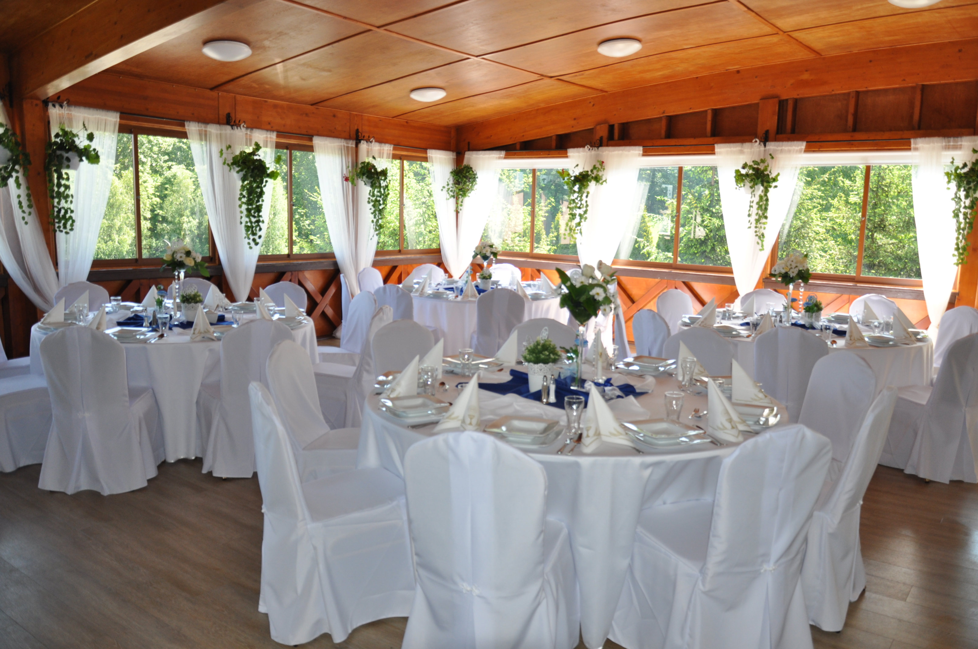 Sala bankietowa z drewnianą konstrukcją, udekorowana białymi obrusami, pokrowcami na krzesła, kwiatami i zwisającymi roślinami przy oknach.