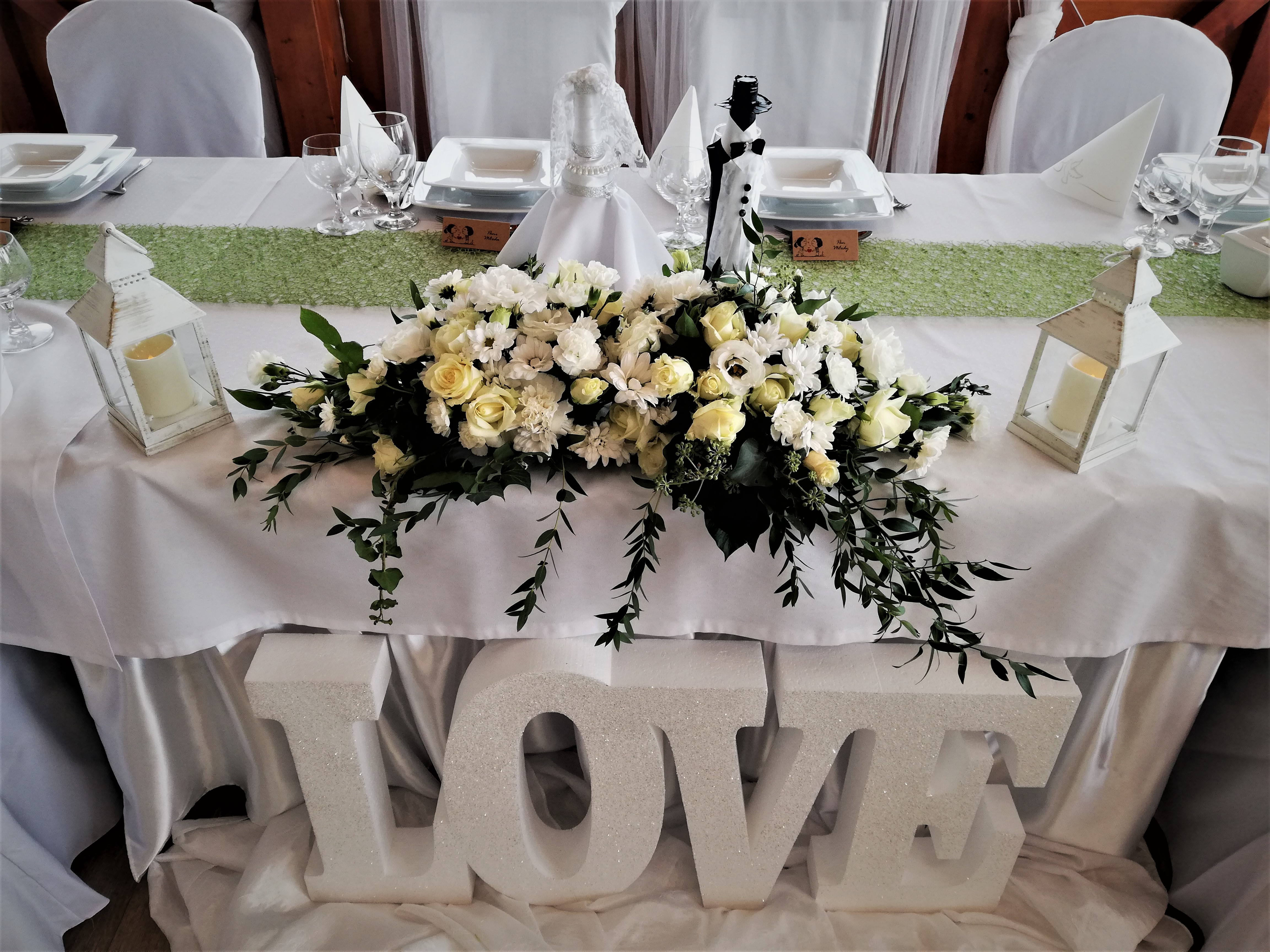 Dekoracja stołu weselnego z białych kwiatów, lampionów i dużych, błyszczących liter LOVE, ustawiona na białym obrusie z zielonym bieżnikiem, widoczne nakrycia dla gości i figurki pary młodej.