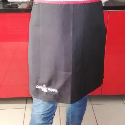 Czarny fartuch kelnerski z haftowanym logo kawiarni Cafe Malina, widoczne niebieskie jeansy i czerwone szafki w tle.