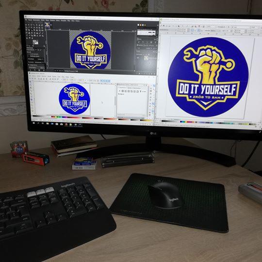 Ekran monitora z otwartym programem graficznym prezentującym projekt graficzny: dłoń trzymająca klucz francuski wpisana w niebieskie koło z napisem 'Do it yourself, Zrób to sam'.