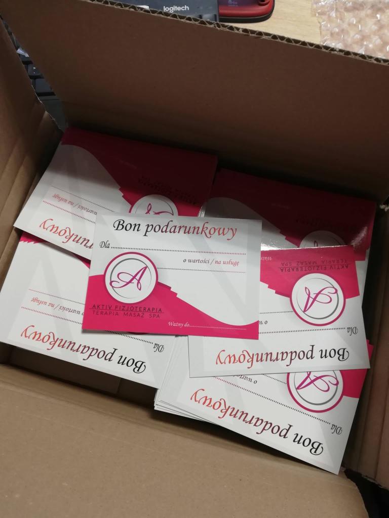 Stos wizytówek z bonami podarunkowymi z logo Aktiv Fizjoterapia Terapia Masaż Spa w kartonowym pudełku, widoczny fragment klawiatury Logitech.
