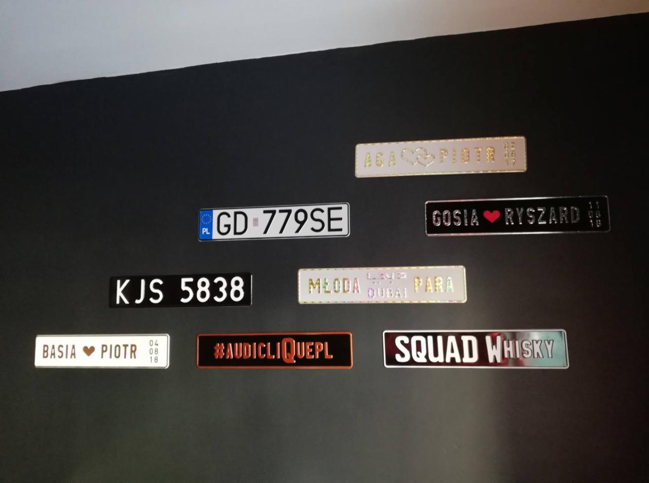 Kolekcja ozdobnych tabliczek z napisami i imionami, imitujących tablice rejestracyjne, umieszczona na ciemnej ścianie.