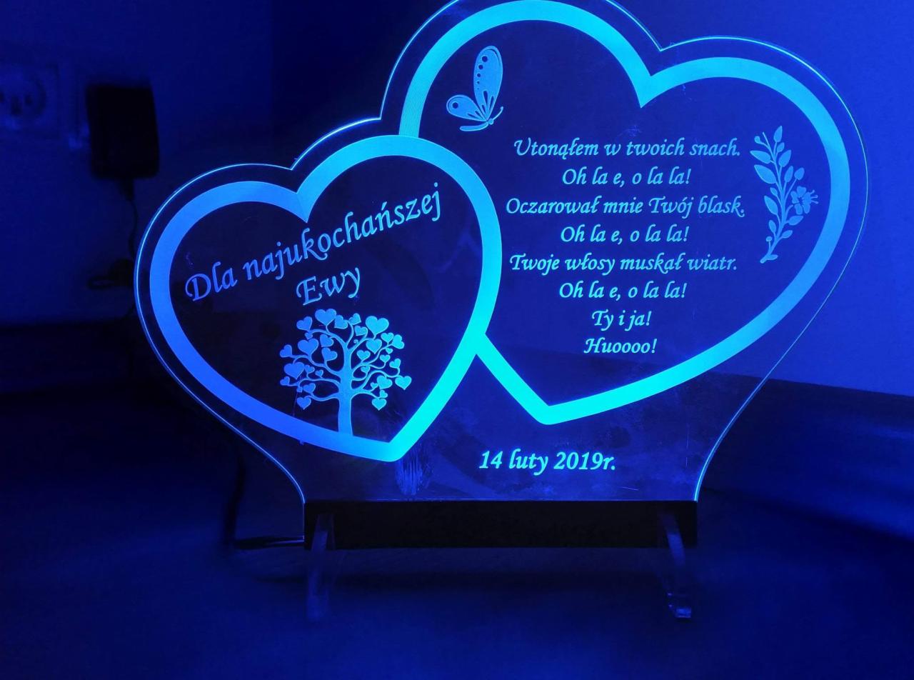 Świecąca, spersonalizowana lampka w kształcie dwóch serc z wygrawerowanym tekstem 'Dla najukochańszej Ewy', wierszem i datą 14 luty 2019r., na przezroczystej podstawce.