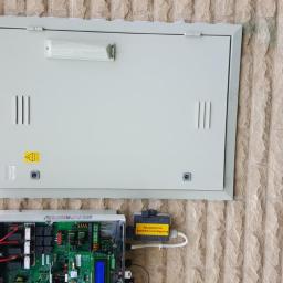 lemar - Szara skrzynka elektryczna zamontowana na teksturowanej ścianie, z otwartym panelem poniżej, odsłaniającym wewnętrzne okablowanie i elektronikę.