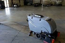 Szara, samojezdna maszyna czyszcząca Magnum na betonowej posadzce w przestronnym, przemysłowym wnętrzu. W tle widoczne pojazdy i słabe oświetlenie.