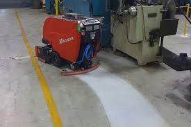 Czerwona maszyna czyszcząca marki Minuteman na betonowej podłodze hali przemysłowej, pozostawiająca za sobą mokry ślad po myciu, widoczna żółta linia na podłodze.