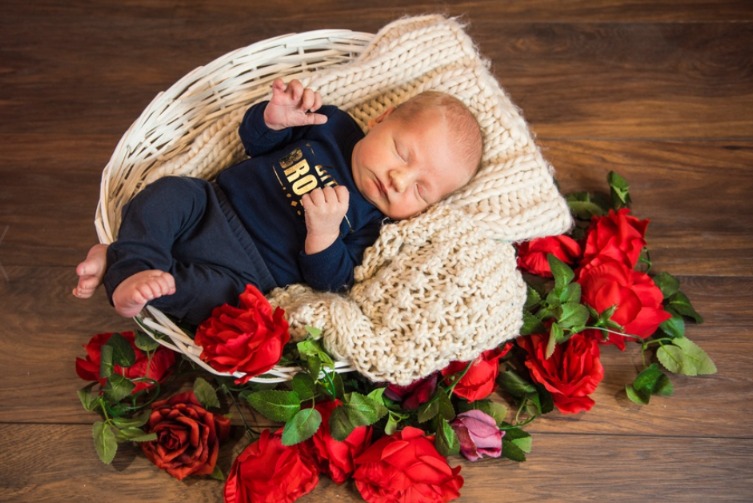 Śpiący noworodek w koszyku wyłożonym kremowym pledem, otoczonym czerwonymi różami na drewnianym tle. Fotografia wykonana w Ostrowcu Świętokrzyskim.