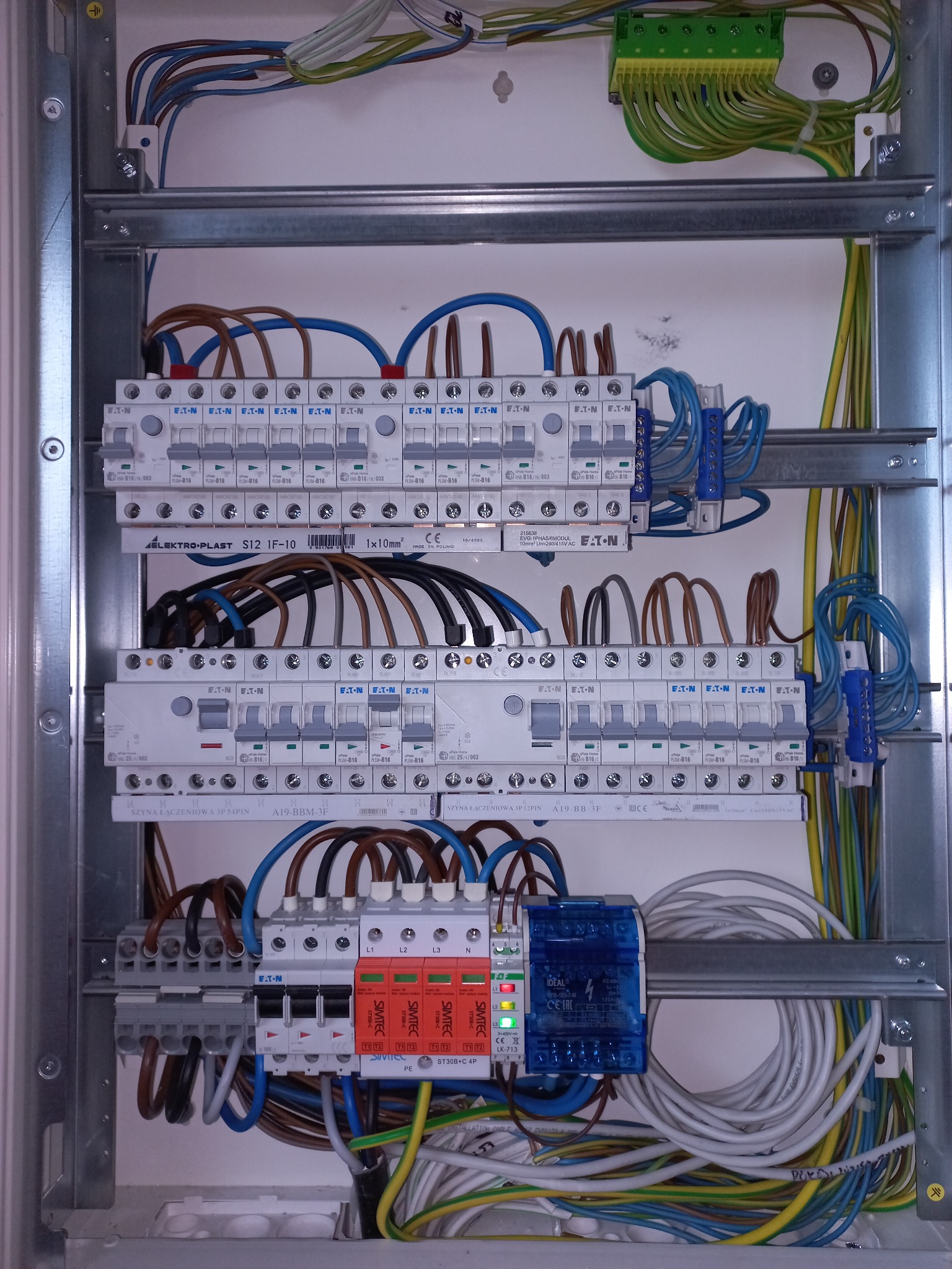 Otwarta skrzynka rozdzielcza z zainstalowanymi bezpiecznikami, ogranicznikami przepięć i okablowaniem w różnych kolorach, widoczne elementy marki Eaton i Simtec.