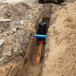 Głęboki wykop w ziemi z widocznymi rurami kanalizacyjnymi: czarną, pomarańczową i niebieską, na tle żółtej maszyny budowlanej i zwiniętych niebieskich rur na powierzchni.