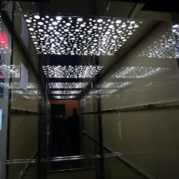 Wnętrze windy z lustrzanymi ścianami i podświetlanym sufitem w geometryczny wzór kropek, odbicie sylwetek osób w głębi kabiny.