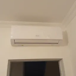 Biały klimatyzator marki Gree zamontowany na ścianie w pomieszczeniu, z folią ochronną na obudowie, widoczny fragment okna poniżej.