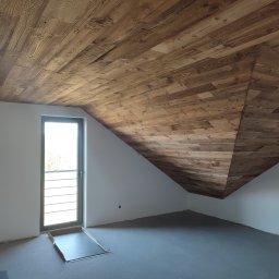 RK ELKO Knopik Rafał - Wykończone drewnem poddasze ze skośnym sufitem i oknem. Widoczne detale drewnianej konstrukcji sufitu i surowa podłoga. Charakterystyczny kształt poddasza.