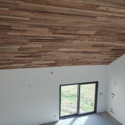 RK ELKO Knopik Rafał - Drewniana boazeria na skośnym suficie w surowym wnętrzu z oknem. Widoczne wystające kable elektryczne. Podłoga betonowa. Naturalne światło dzienne.