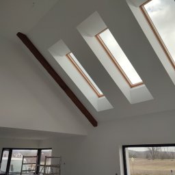 RK ELKO Knopik Rafał - Poddasze po malowaniu na biało z trzema oknami dachowymi i drewnianą belką stropową. Widoczne rusztowania i okno z widokiem na zewnątrz.