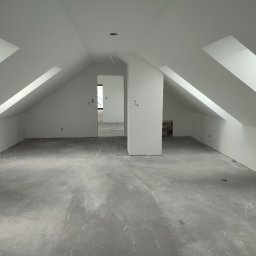 Knopik Rafał Lipusz - Poddasze po remoncie, świeżo pomalowane na biało, z widocznymi skosami i oknami dachowymi. Surowa podłoga, przygotowana do dalszych prac wykończeniowych. Widok na kolejne pomieszczenia.