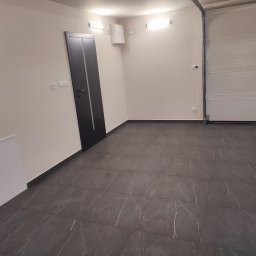 Knopik Rafał Lipusz - Nowo wykończone wnętrze z ciemnoszarymi płytkami podłogowymi z białymi żyłkami, beżowymi ścianami i nowoczesnymi drzwiami. Widoczna biała brama garażowa i skrzynka elektryczna.