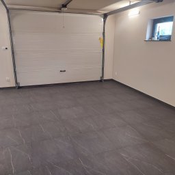 Knopik Rafał Lipusz - Wnętrze garażu z białą bramą segmentową, ciemnoszarymi płytkami podłogowymi z białym żyłkowaniem i beżowymi ścianami. Na parapecie okna stoją tuby z silikonem.
