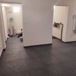 Knopik Rafał Lipusz - Nowocześnie wykończony korytarz z ciemnymi płytkami imitującymi marmur i beżowymi ścianami. W tle widoczny system filtracji wody oraz narzędzia remontowe.